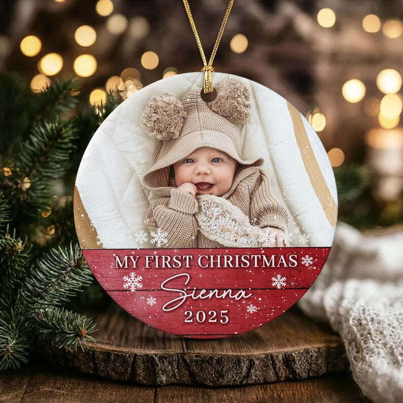 A round ceramic Christmas ornament featuring a photograph of a baby in a beige knit hat with pom-poms. The bottom section has a red wood-grain background with white snowflakes and the text: MY FIRST CHRISTMAS Sienna 2025.
