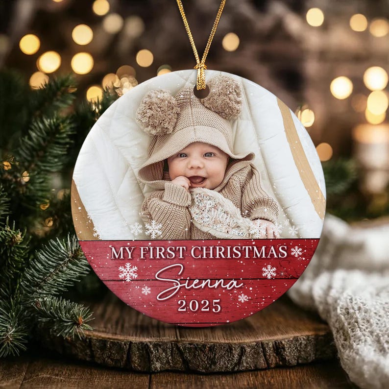 A round ceramic Christmas ornament featuring a photograph of a baby in a beige knit hat with pom-poms. The bottom section has a red wood-grain background with white snowflakes and the text: MY FIRST CHRISTMAS Sienna 2025.
