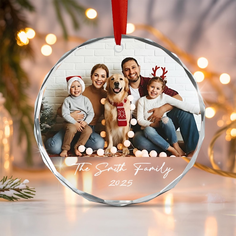 A circular glass Christmas ornament featuring a printed photograph of a family with a dog. The image shows two adults, two children, and a golden retriever wearing a red scarf. The text The Smith Family 2025 is printed in white.