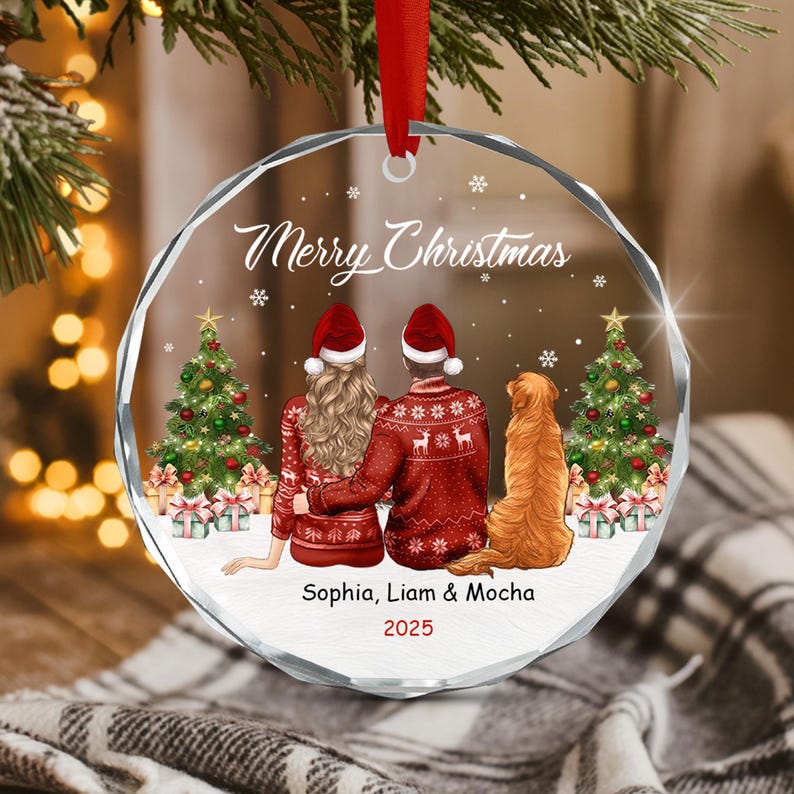 A round, clear acrylic Christmas ornament featuring a printed illustration of two people and a golden retriever dog sitting together. The text reads Merry Christmas, Sophia, Liam & Mocha, and 2025. It hangs from a red ribbon.