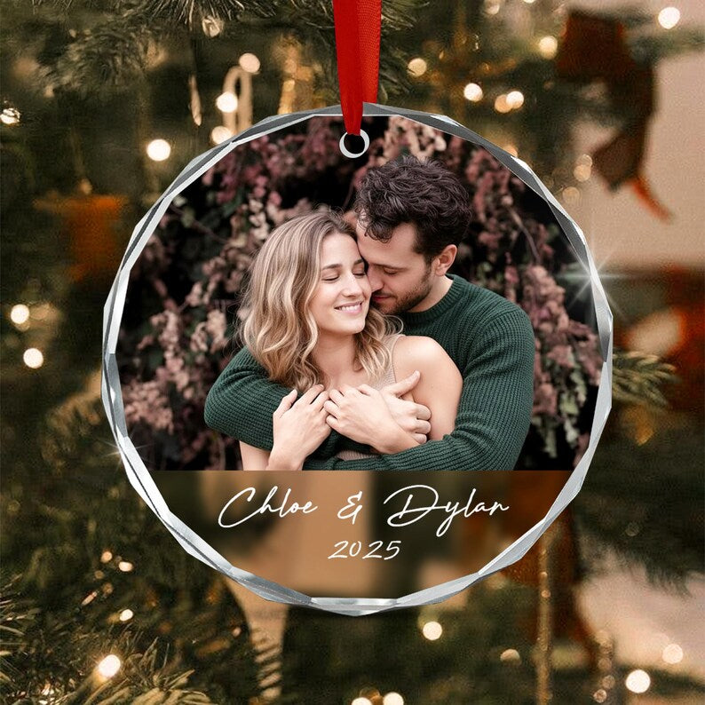A circular glass ornament with faceted edges featuring a photograph of two people embracing. Below the photo, the text Chloe & Dylan and the year 2025 are printed in white script. A red ribbon is attached to the top for hanging.