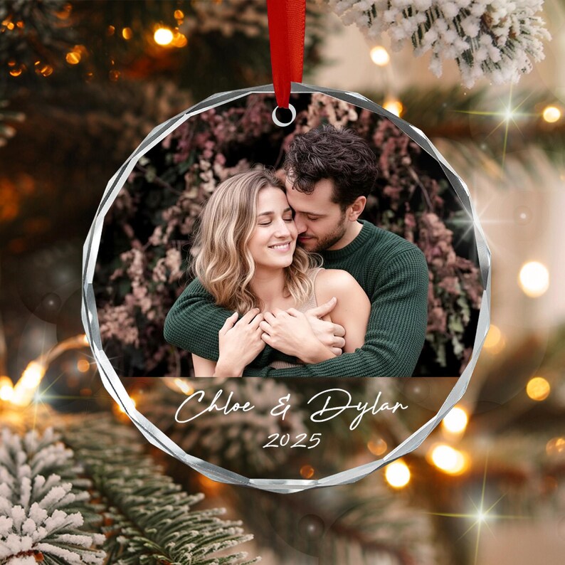 A round, faceted glass ornament featuring a photograph of a couple embracing. The names Chloe & Dylan and the year 2025 are printed in white script below the photo. A red ribbon is attached to the top for hanging on a tree.