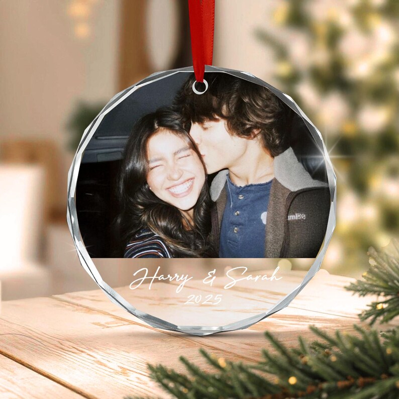 A circular glass ornament featuring a photograph of two people, with the text Harry & Sarah and the year 2025 printed in white script below the image. The ornament has a faceted edge and hangs from a red ribbon.