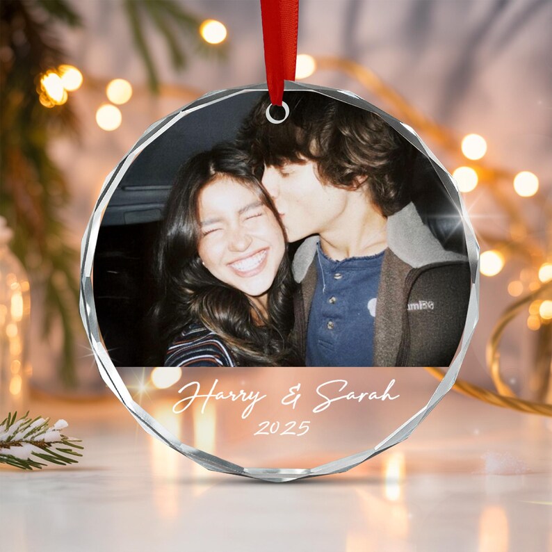 A circular glass ornament featuring a photograph of two people, with one person kissing the other on the cheek. Below the photo, the text reads Harry & Sarah 2025 in white script.