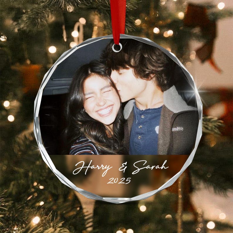 A round, faceted glass Christmas ornament featuring a photograph of two people, with the printed text Harry & Sarah and the year 2025 below the image. The ornament is suspended by a red ribbon and has a clear, reflective edge.