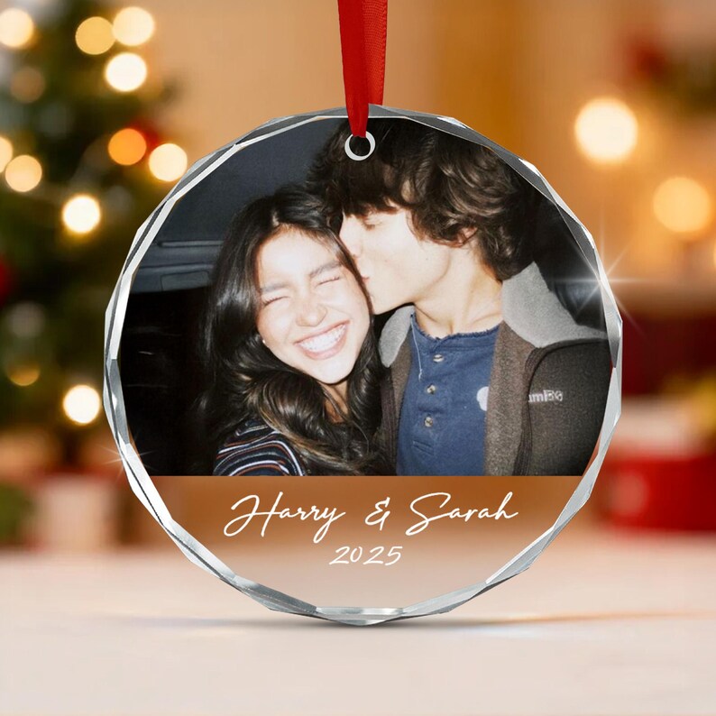 A circular, faceted glass ornament featuring a black and white photograph of two people, with the text Harry & Sarah and 2025 printed in white on a brown background at the bottom, suspended by a red ribbon for hanging.