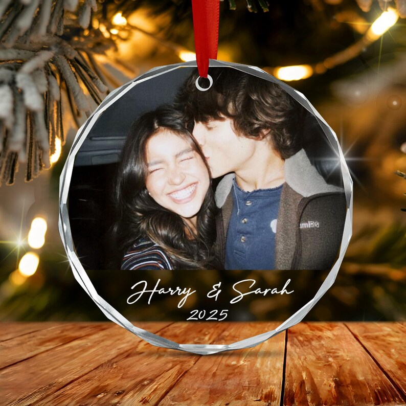 A circular glass ornament featuring a photograph of two people, with the text Harry & Sarah and the year 2025 printed in white script below the image. The ornament is suspended by a red ribbon and has a faceted, clear glass edge.