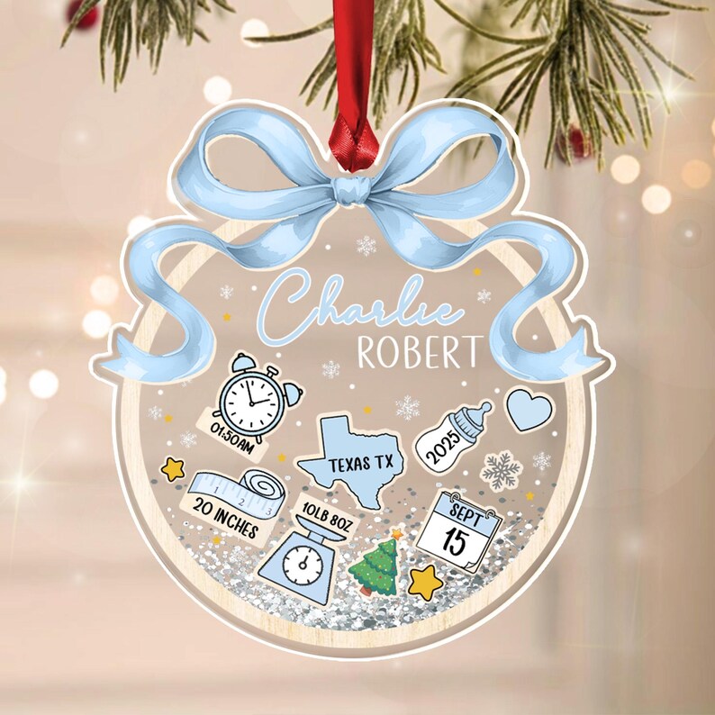 A personalized Christmas ornament with a light blue bow and red ribbon. The ornament features the name "Charlie Robert" and includes baby-related icons, measurements of 20 inches and 10lb 8oz, and the date September 15.