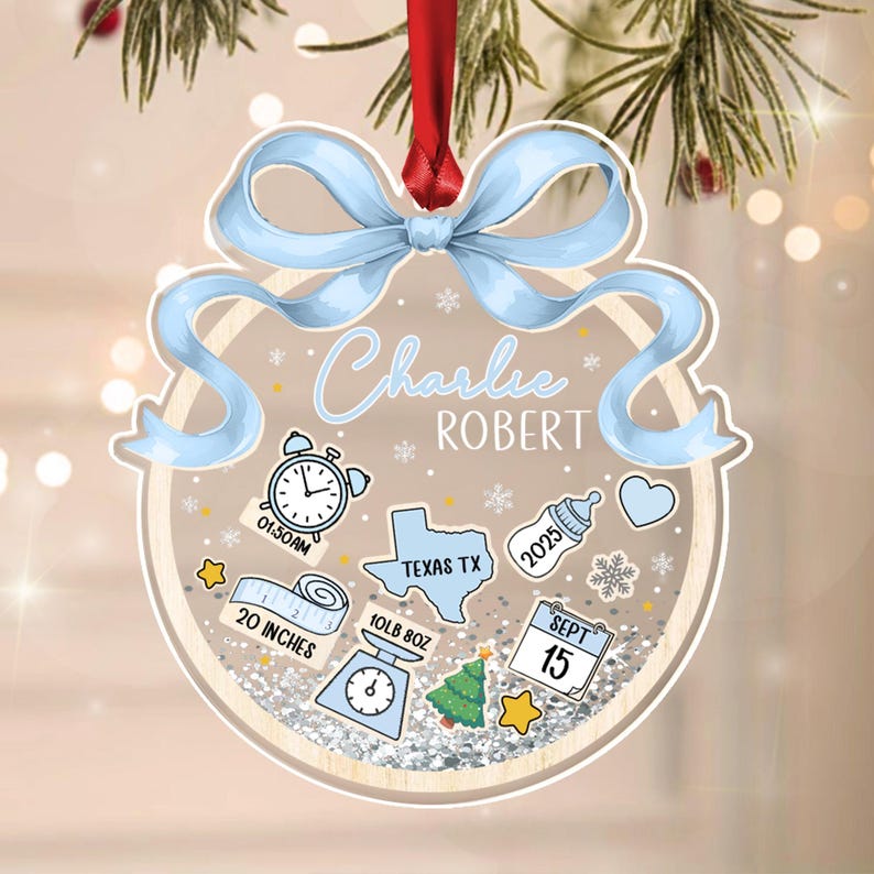 A personalized Christmas ornament with a light blue bow and red ribbon. The ornament features the name "Charlie Robert" and includes baby-related icons, measurements of 20 inches and 10lb 8oz, and the date September 15.