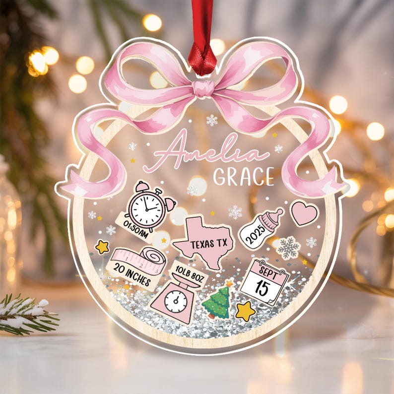 A personalized baby ornament with a pink bow and the name "Amelia Grace." The ornament includes illustrations of a clock, a Texas outline, a baby bottle, a heart, a measuring tape (20 inches), a scale (10lb 8oz), a Christmas tree.