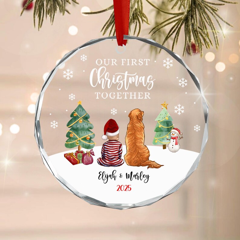 A circular clear glass Christmas ornament with a red ribbon hanger. The design features a child in red and white striped pajamas and a golden dog sitting between two Christmas trees and a snowman.