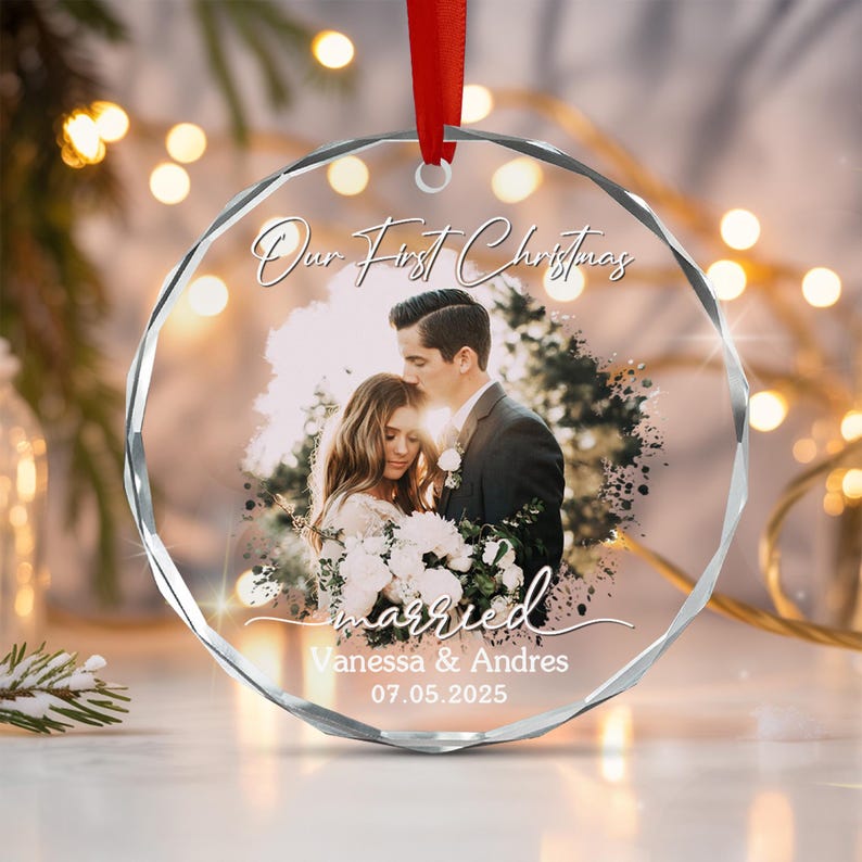 A circular, faceted clear glass ornament featuring a red ribbon hanger. The center displays a photograph of a couple, with the printed text Our First Christmas, married, Vanessa & Andres, and the date 07.05.2025 in white script.