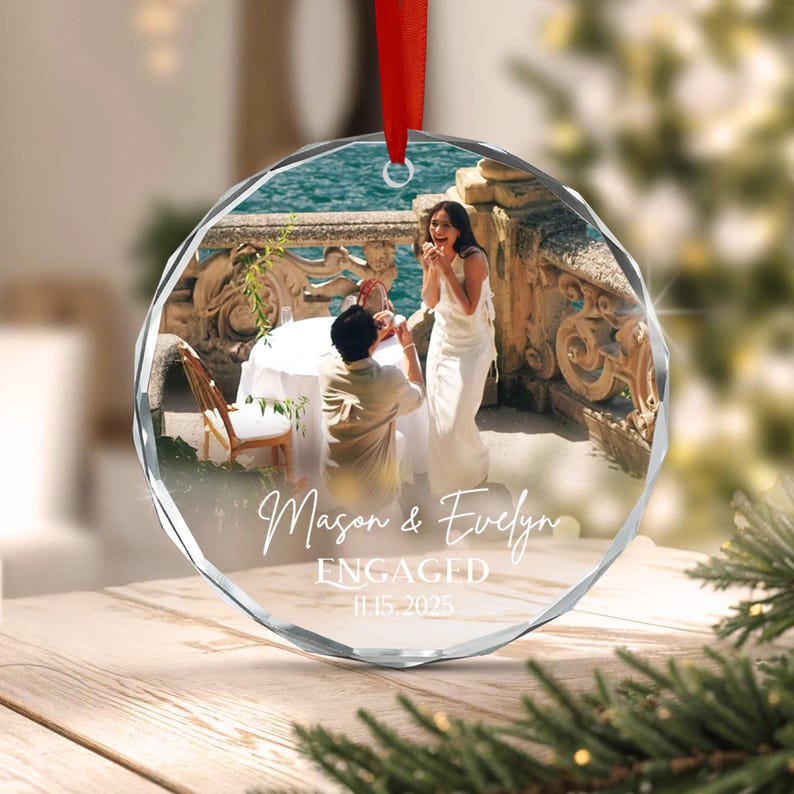 A round, faceted glass ornament featuring a printed photograph of a marriage proposal on a terrace. The text reads Mason & Evelyn ENGAGED 11.15.2025. The ornament is suspended by a red ribbon and displays a couple near the water.