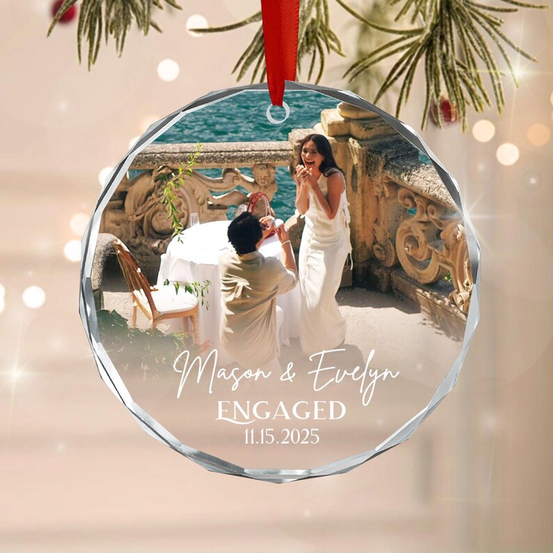 A circular crystal ornament featuring a photograph of a couple during an engagement proposal. The text Mason & Evelyn ENGAGED 11.15.2025 is printed in white across the center.