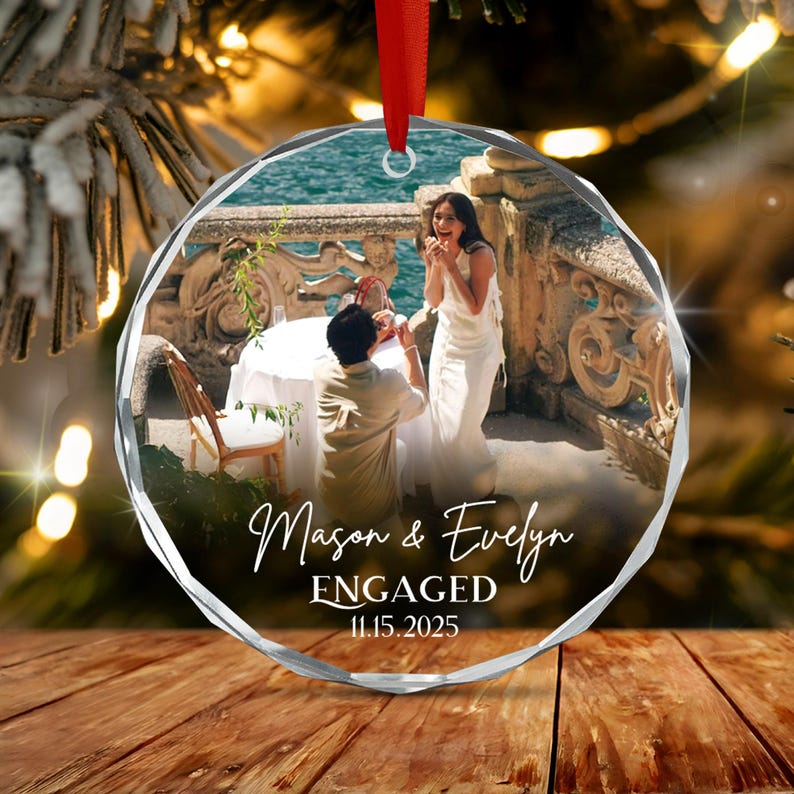 A round, faceted glass ornament featuring a printed photograph of a couple during a proposal on a terrace. The text Mason & Evelyn ENGAGED 11.15.2025 is printed in white script and serif fonts.