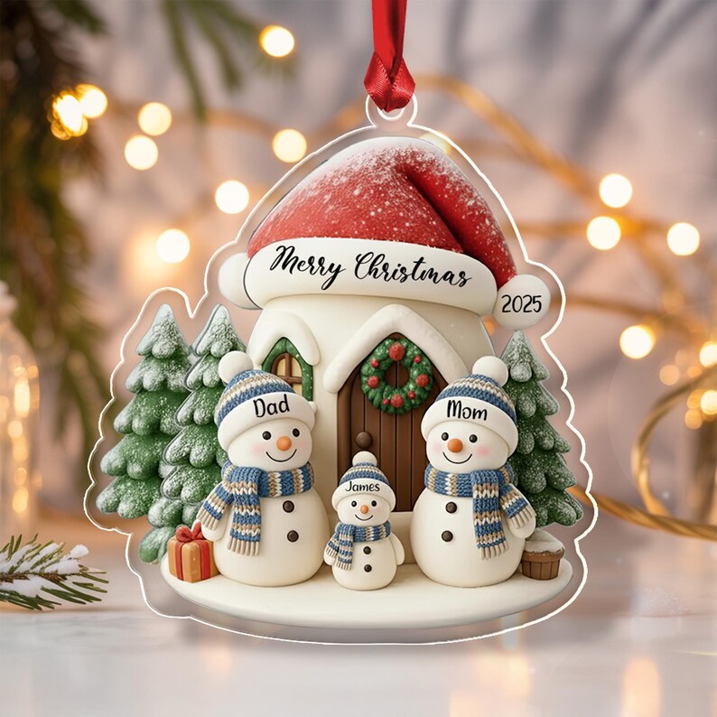 A clear acrylic Christmas ornament shaped like a house with a red Santa hat roof. It features three snowmen labeled Dad, Mom, and James, wearing blue and white striped hats and scarves.