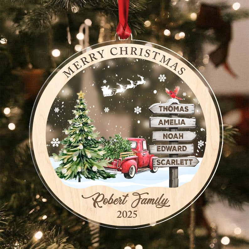 A round, clear acrylic Christmas ornament with a wooden border, featuring a red truck, a green Christmas tree, a signpost with the names Thomas, Amelia, Noah, Edward, and Scarlett, a Santa sleigh silhouette.