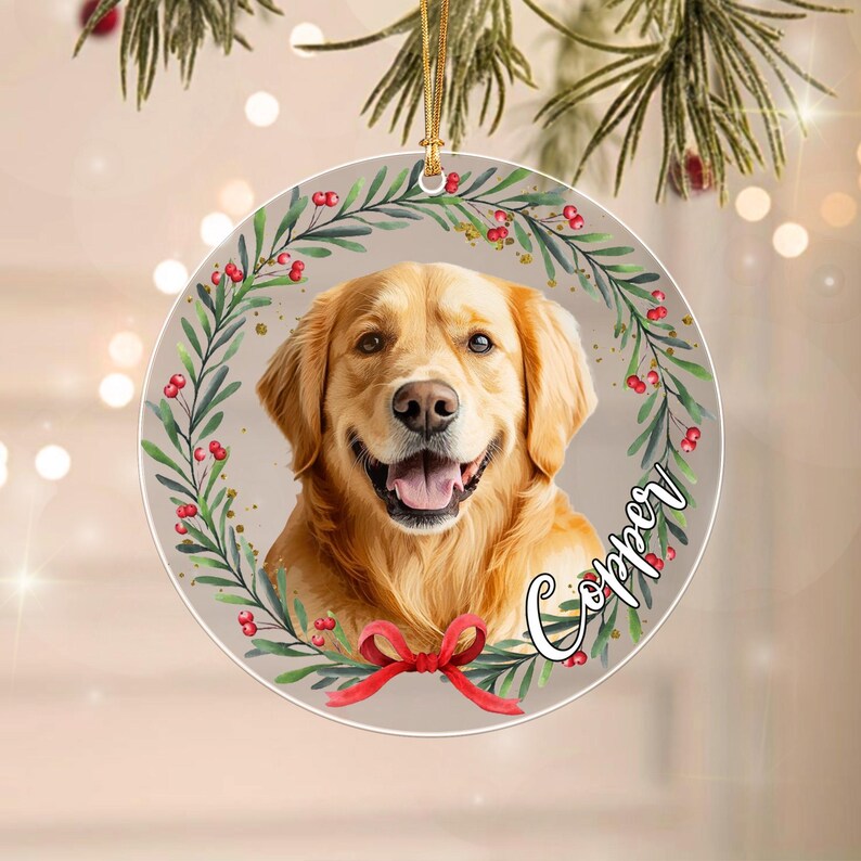 A round ceramic ornament featuring a golden retriever portrait centered within a green leafy wreath with red berries. A red ribbon is tied at the bottom of the wreath. The name Copper is written in black script on the right side.