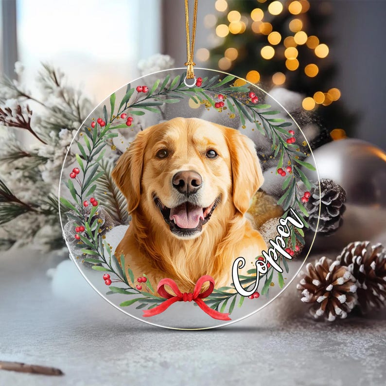 A round, clear acrylic Christmas ornament featuring a portrait of a golden retriever dog. The dog is framed by a green wreath with red berries and a red ribbon at the bottom.