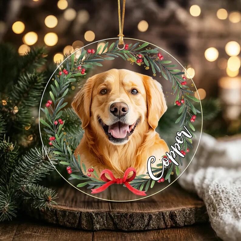 A circular clear acrylic ornament featuring a portrait of a golden retriever dog centered within a wreath of green leaves and red berries. A red ribbon bow is at the bottom, and the name Copper is written in white script text.