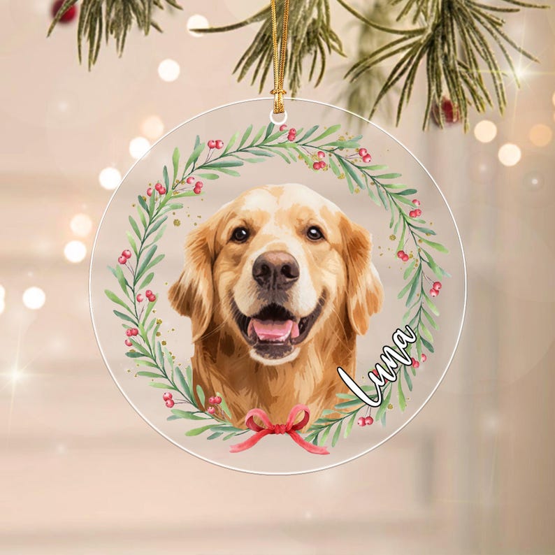 A circular clear acrylic ornament featuring a golden retriever portrait centered within a green leafy wreath with red berries. A red ribbon bow is at the bottom of the wreath.