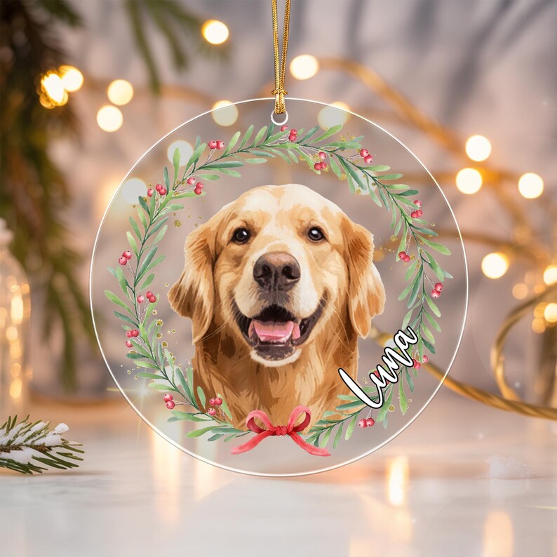 A circular clear acrylic ornament featuring a portrait of a golden retriever dog centered within a wreath of green leaves and red berries.