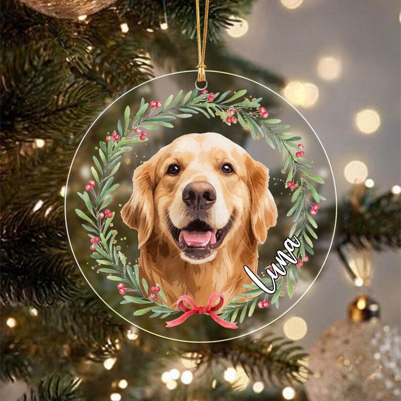A round, clear acrylic Christmas ornament featuring a portrait of a golden retriever dog centered within a wreath of green leaves and red berries. A red ribbon bow is at the bottom, and the name Luna is written in white script.