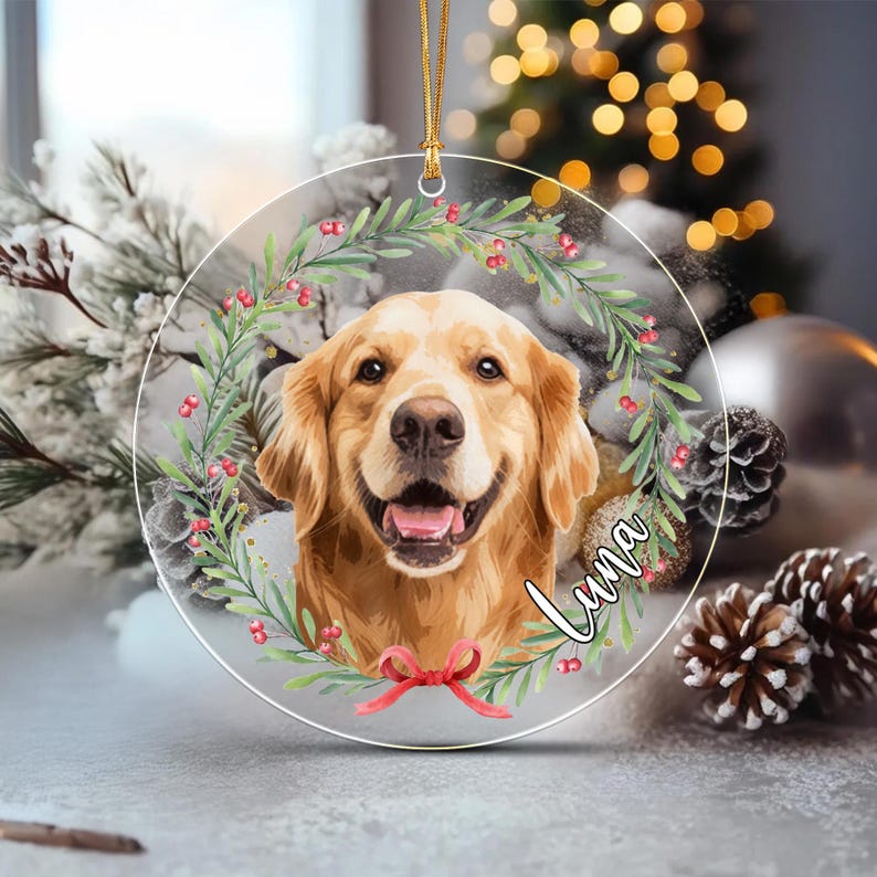 A round, clear acrylic ornament featuring a golden retriever portrait centered within a green wreath adorned with red berries and a red bow at the bottom.