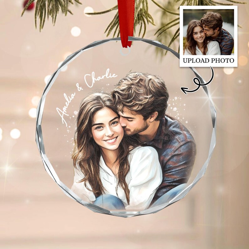 A circular clear glass ornament featuring a custom illustration of a couple embracing. The text Amelia & Charlie is printed in white script. A red ribbon is attached to the top for hanging. The design is based on a provided photo.