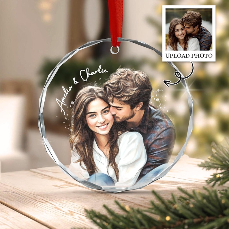 A circular, faceted glass ornament featuring a printed illustration of a couple embracing. The text 'Amelia & Charlie' is written in white script on the left side. A red ribbon is attached to the top for hanging purposes.
