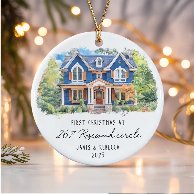 A round white ceramic ornament featuring a watercolor illustration of a blue two-story house with brown shutters. The text reads: FIRST CHRISTMAS AT 267 Rosewood circle JAVIS & REBECCA 2025.