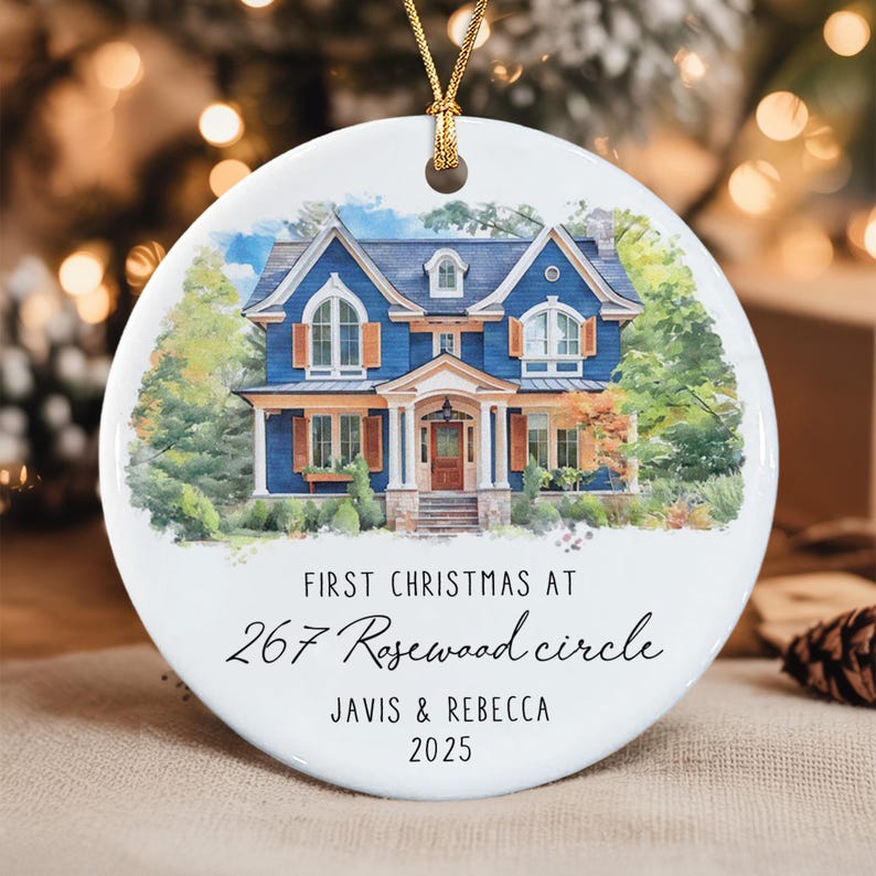 A round white ceramic ornament featuring a watercolor illustration of a blue two-story house with brown shutters. The text reads: FIRST CHRISTMAS AT 267 Rosewood circle JAVIS & REBECCA 2025.