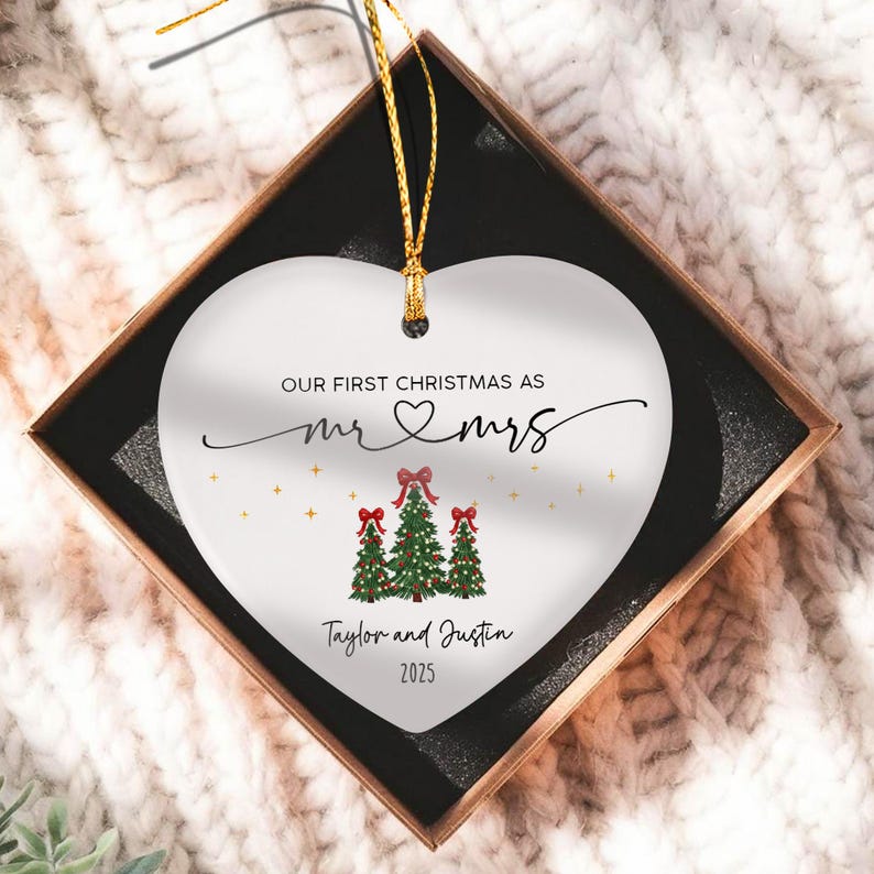 A white heart-shaped ceramic ornament with a gold hanging string. The text reads "OUR FIRST CHRISTMAS AS mr & mrs" above three illustrated green Christmas trees with red bows, followed by the names "Taylor and Justin" and "2025".