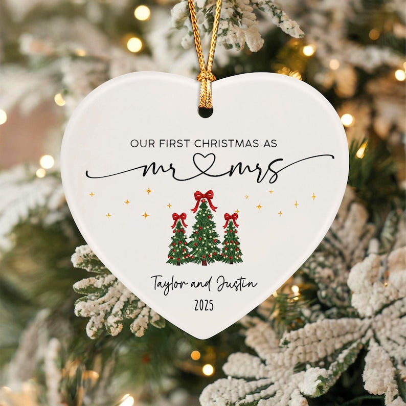 A white heart-shaped ceramic ornament featuring the text "OUR FIRST CHRISTMAS AS mr & mrs" with a heart symbol, three green Christmas trees with red bows, the names "Taylor and Justin," and the year "2025" in black and gold.
