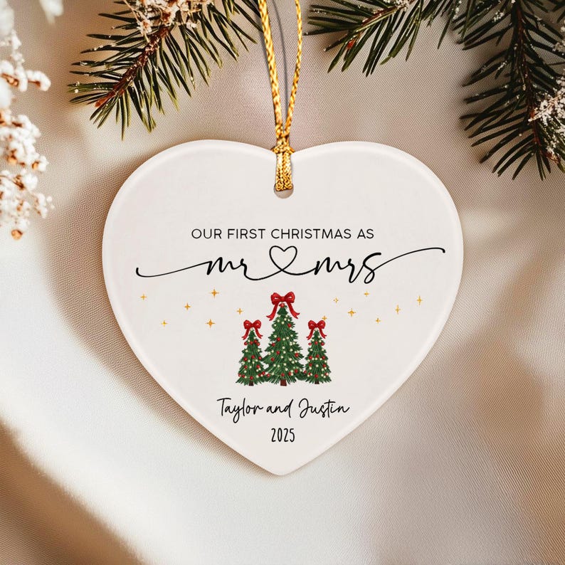 A white heart-shaped ceramic ornament hanging from a gold string. It features the text "OUR FIRST CHRISTMAS AS mr & mrs", "Taylor and Justin", and "2025".