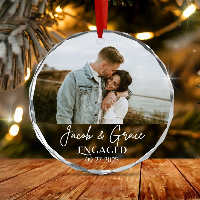 A circular, faceted glass ornament featuring a photograph of a couple embracing outdoors. The text "Jacob & Grace", "ENGAGED", and "09.27.2025" is printed in white script and block lettering. A red ribbon is attached to the top.