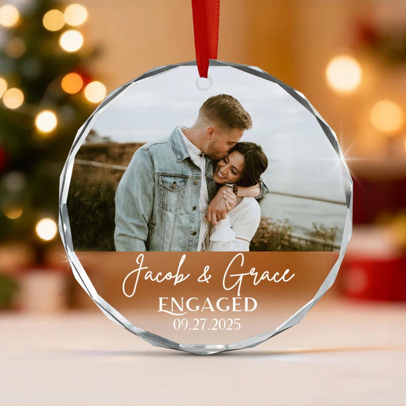A circular glass ornament with a faceted edge, featuring a photograph of a couple embracing outdoors. Below the photo, the text reads Jacob & Grace, ENGAGED, and 09.27.2025 in white script and block lettering.
