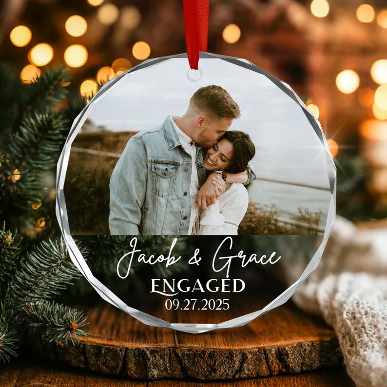 A circular glass ornament featuring a photograph of a couple embracing. The text reads Jacob & Grace ENGAGED 09.27.2025.