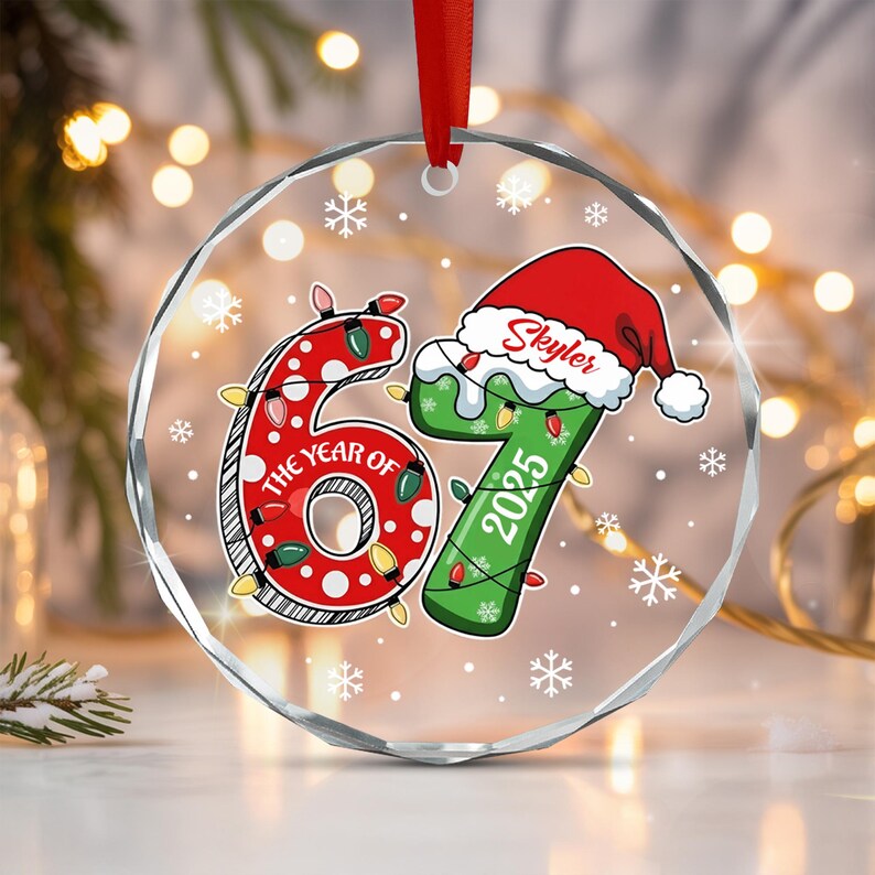 A circular clear acrylic ornament featuring the number 67, with the red number 6 decorated with white dots and the green number 7 wearing a Santa hat with the name Skyler. It includes the text THE YEAR OF 2025 and string lights.