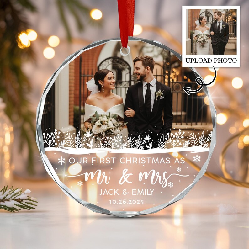 A circular glass ornament featuring a printed wedding photograph of a couple, white floral line art, and the text "OUR FIRST CHRISTMAS AS MR & MRS JACK & EMILY 10.26.2025".