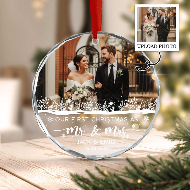A round, clear acrylic Christmas ornament featuring a custom wedding photograph of a couple. The ornament includes white printed text that reads "OUR FIRST CHRISTMAS AS MR & MRS JACK & EMILY 10.26.2025" with a red ribbon hanger.