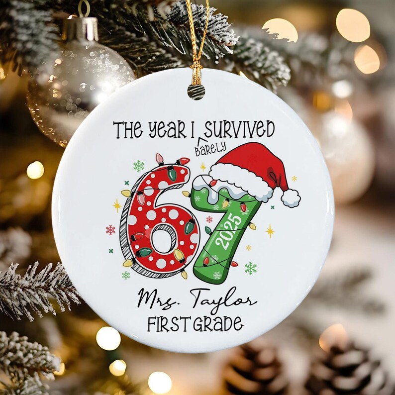 A round white ceramic ornament featuring the text THE YEAR I SURVIVED BARELY, the number 67 with red polka dots and green coloring, a Santa hat, the year 2025, and the text Mrs. Taylor FIRST GRADE, all hanging by a gold string.