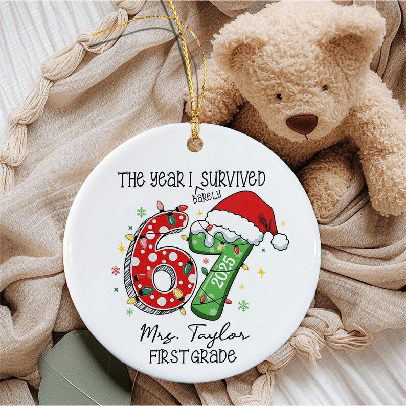 A round white ceramic ornament with a gold hanging string. The text reads THE YEAR I BARELY SURVIVED 67, 2025, Mrs. Taylor, and FIRST GRADE. The number 67 is decorated with red and green Christmas lights and a red Santa hat.