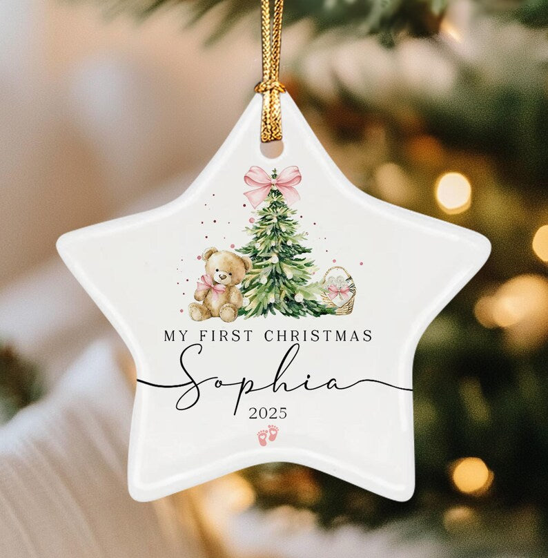 A white star-shaped ceramic ornament with a gold hanging cord. The design features a watercolor teddy bear, a Christmas tree with a pink bow, and the text "MY FIRST CHRISTMAS Sophia 2025" with two small pink baby footprints below.