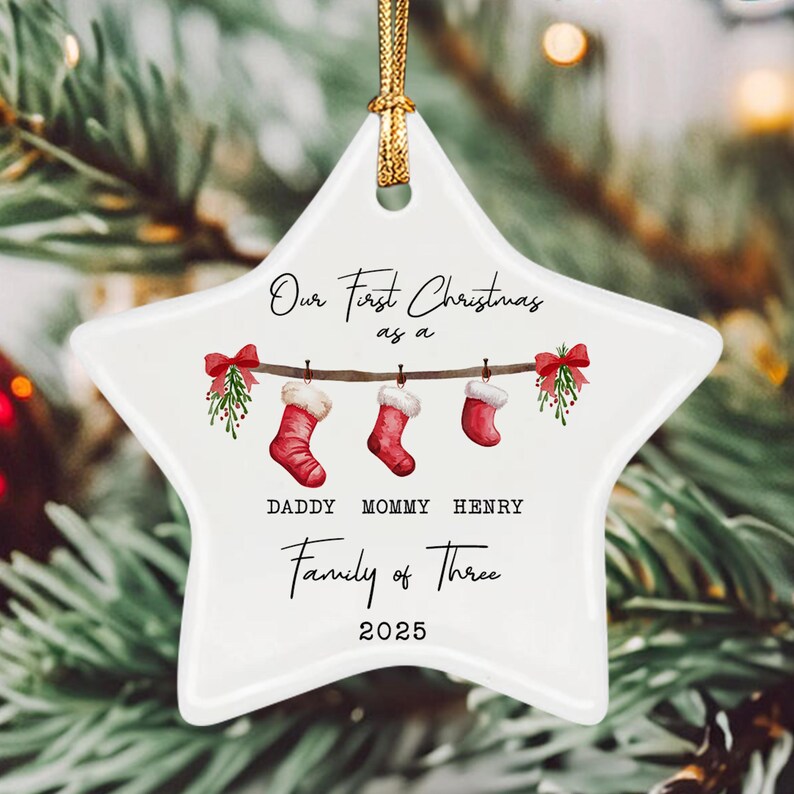 A white star-shaped ceramic ornament with a gold hanging cord. The design features three red Christmas stockings hanging from a branch with mistletoe, text reading Our First Christmas as a DADDY MOMMY HENRY Family of Three 2025.