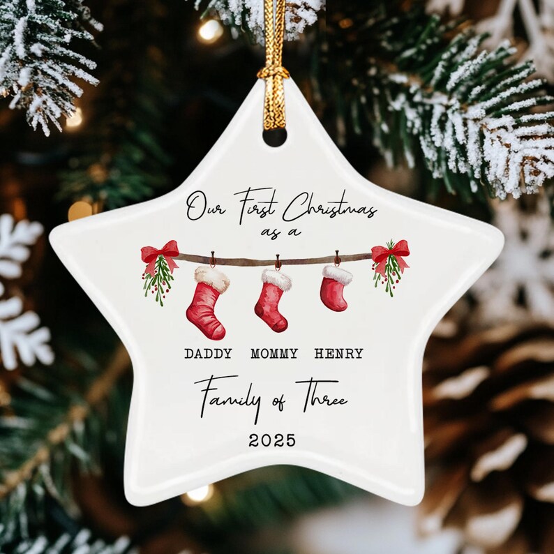 A white star-shaped ceramic ornament featuring a gold hanging cord. The design includes three red stockings hanging from a branch with mistletoe, text reading Our First Christmas as a, DADDY MOMMY HENRY, Family of Three, and 2025.