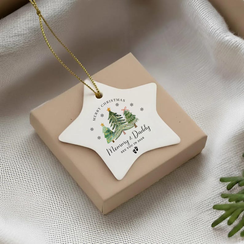 A white star-shaped ceramic ornament with a gold hanging string. The design features watercolor Christmas trees, snowflakes, and the text Merry Christmas Mommy & Daddy See You In 2026 with two small footprint icons below the text.