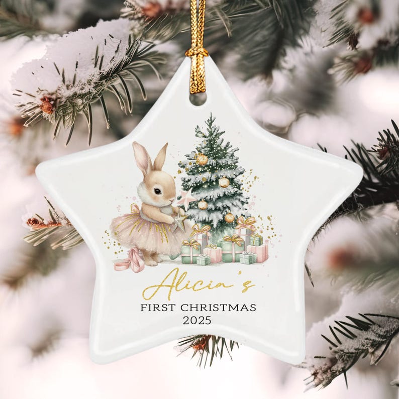 A white star-shaped ceramic ornament featuring a watercolor illustration of a rabbit in a tutu standing next to a decorated Christmas tree and gifts. The text reads Alicia's FIRST CHRISTMAS 2025 in gold and black lettering.