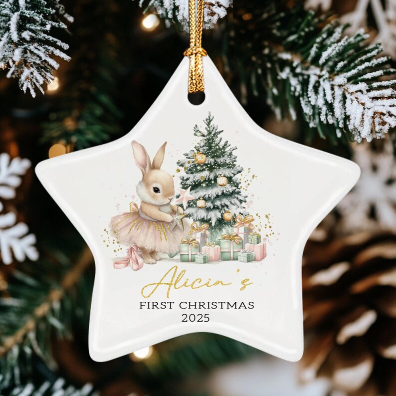 A white star-shaped ceramic ornament with a gold hanging cord. The design features a rabbit in a pink tutu next to a decorated Christmas tree and gifts. Text reads: Alicia's FIRST CHRISTMAS 2025. The surface is smooth and glossy.