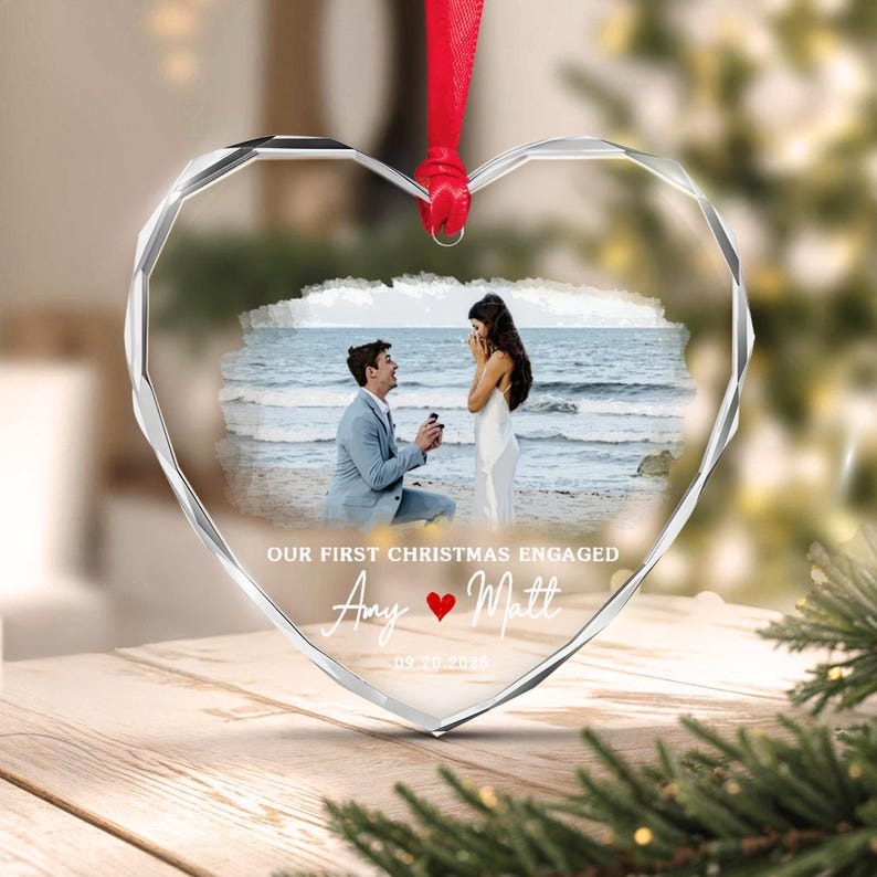 A heart-shaped clear acrylic ornament featuring a printed photograph of a marriage proposal on a beach. The text reads OUR FIRST CHRISTMAS ENGAGED, Amy, Matt, and 09.20.2025, with a red heart icon. It hangs from a red ribbon.